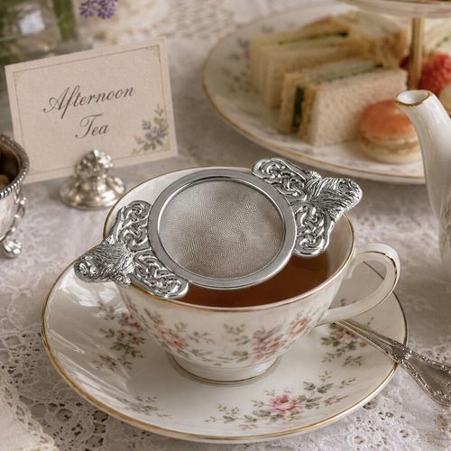 A stainless steel mesh tea strainer with decorative pewter handles featuring a highland cow head and intricate Celtic knotwork, resting across a floral porcelain teacup during an afternoon tea service