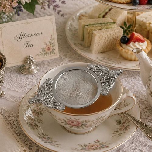 A stainless steel mesh tea strainer with ornate pewter handles featuring a highland stag head and Celtic knotwork, resting across a floral porcelain teacup during an afternoon tea service