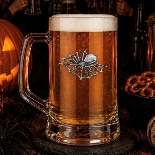 A clear glass beer tankard with a sturdy handle, filled with amber lager and a thick white foam head, featuring a detailed pewter emblem of a spider sitting on an intricate spiderweb