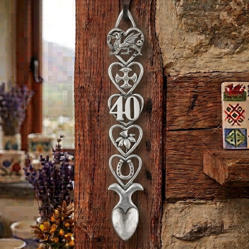 Handcrafted pewter 40th birthday Welsh lovespoon hanging on a rustic wooden beam, featuring detailed symbols including a Welsh dragon, Celtic cross, the number 40, a daffodil, a horseshoe, and a heart-shaped spoon bowl.