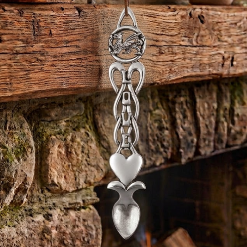 Handcrafted pewter Welsh lovespoon hanging against a wooden beam and stone fireplace, featuring a circular Welsh dragon top, the word "LOVE" carved vertically through open Celtic knotwork, a smooth solid heart, and a spoon bowl.