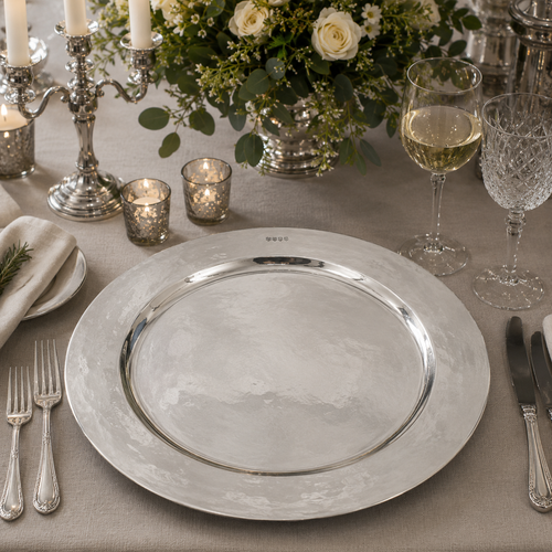 Handcrafted pewter 19 inch charger plate with polished hammered finish, luxury dining table centrepiece displayed with crystal glassware, silver cutlery, candlelight and elegant floral arrangement