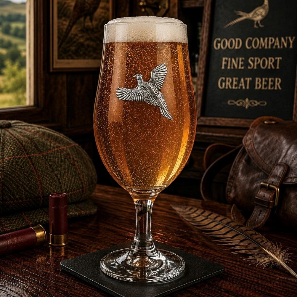 A stemmed beer chalice glass filled with golden ale and a frothy white head, featuring a raised pewter emblem of a pheasant bird in mid-flight