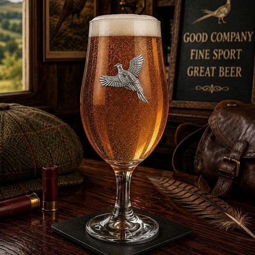 A stemmed beer chalice glass filled with golden ale and a frothy white head, featuring a raised pewter emblem of a pheasant bird in mid-flight