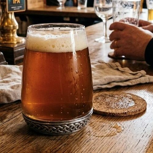 A curved glass drinks tumbler filled with amber ale and a thick white foam head, featuring a heavy pewter base detailed with an intricate interwoven Celtic knotwork band
