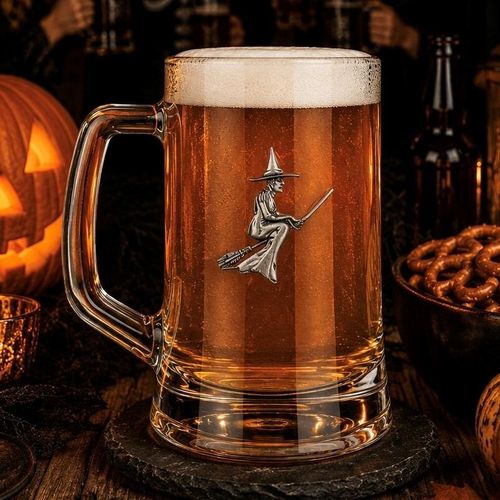 A clear glass beer tankard with a sturdy handle, filled with amber lager and a thick white foam head, featuring a detailed pewter emblem of a witch flying on a broomstick