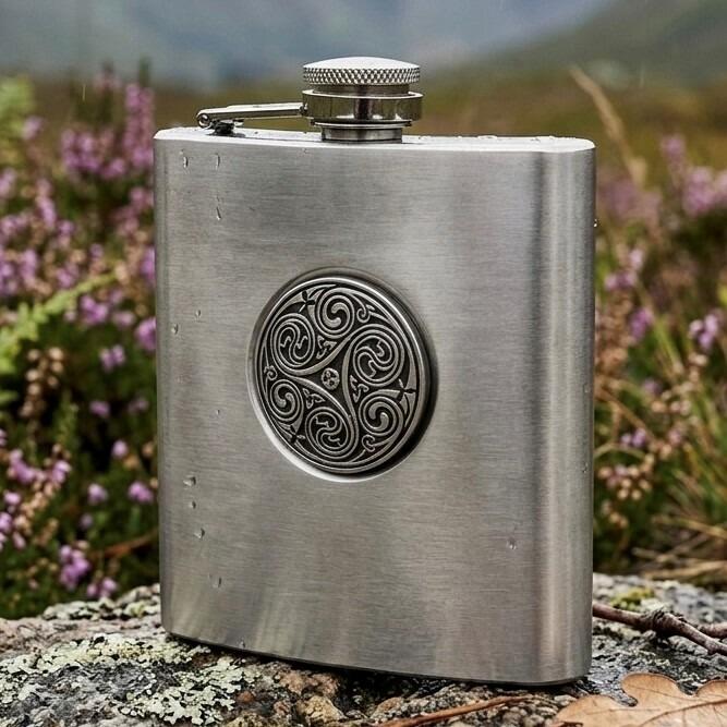 Brushed stainless steel hip flask featuring a central circular pewter medallion with an intricate Celtic triskele triple spiral, displayed outdoors on a wet stone with purple heather in the background