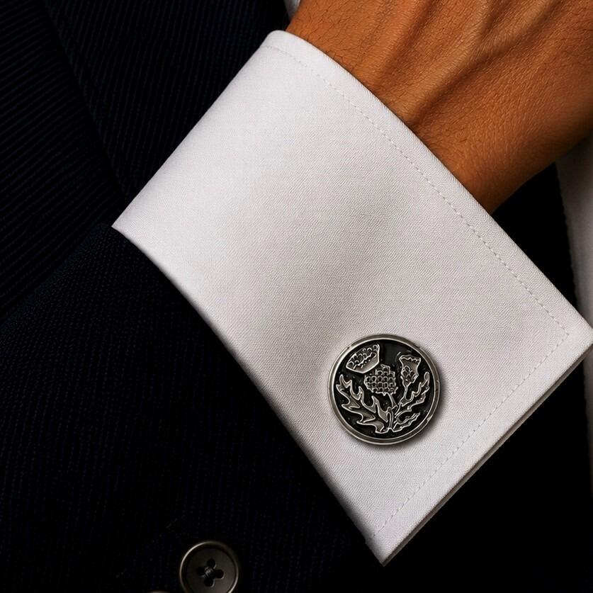 A circular pewter cufflink featuring an embossed Scottish thistle design with a textured background and polished borders