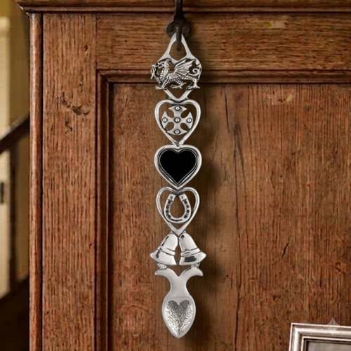 Handcrafted pewter Welsh lovespoon hanging on a wrought iron hook against a wooden panelled wall, featuring a spoked wheel top, a heart frame with a central black heart insert, a lucky horseshoe, twin wedding bells, and a textured spoon bowl.