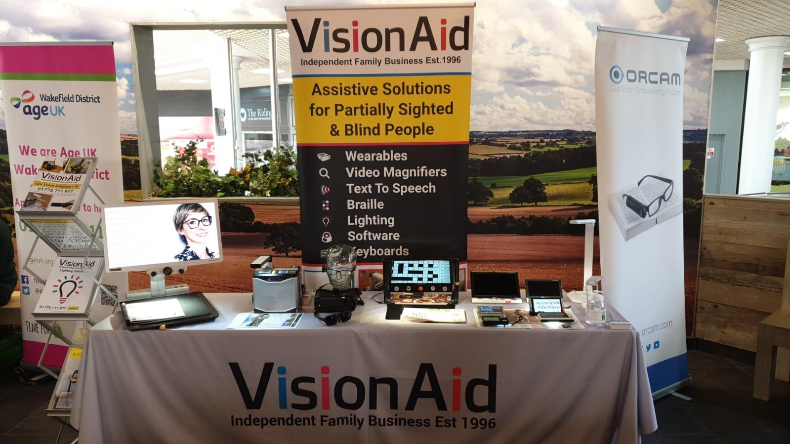 Image shows a table with a selection of our low vision and blindness solutions behind the table there is a visionaid and orcam banner to th eleft side of the table is a stand full of our brochures