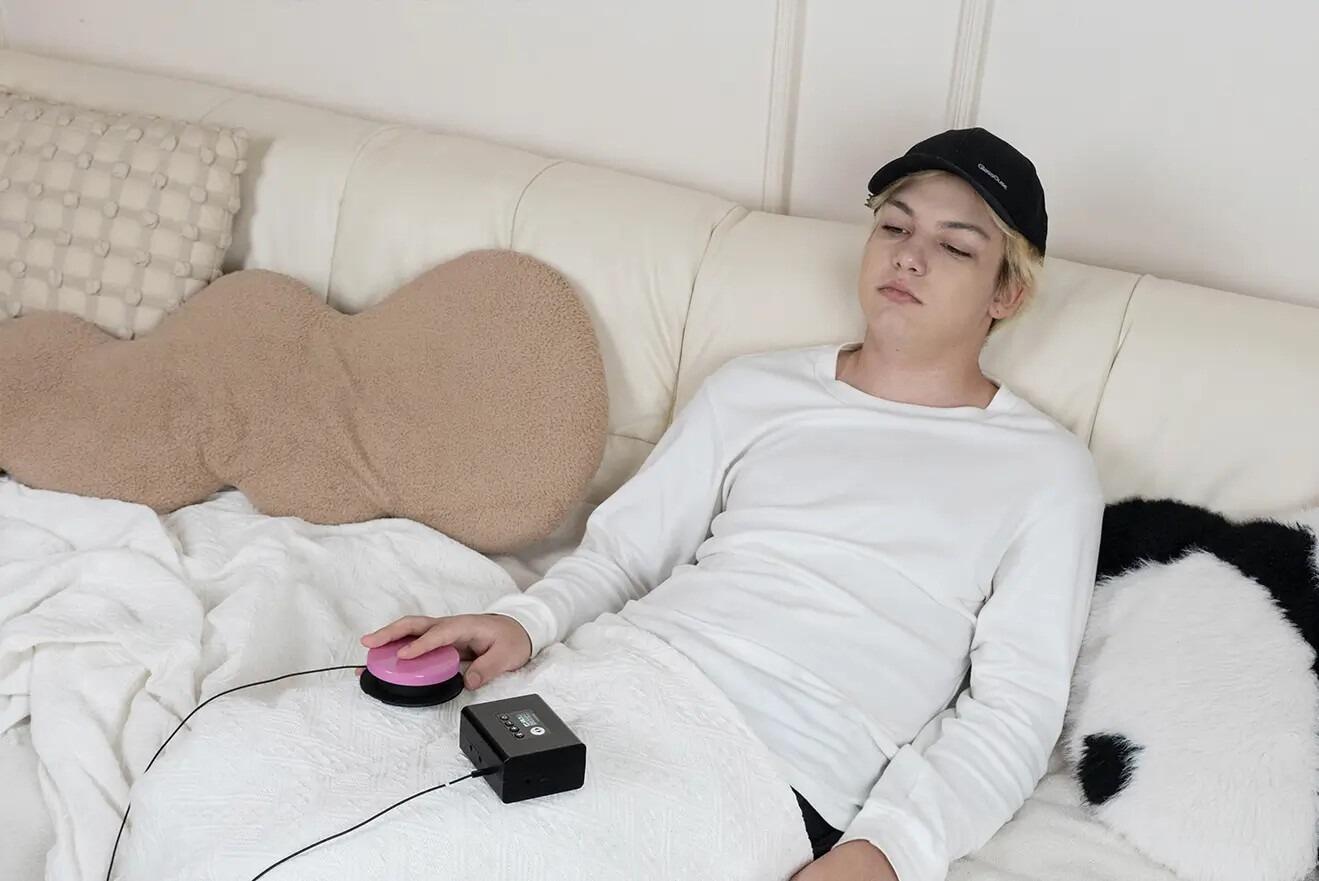 A young man on a sofa with the GlassOuse G-Speech on his lap, there is a press switch attached to the G-Speech and he is pressing it