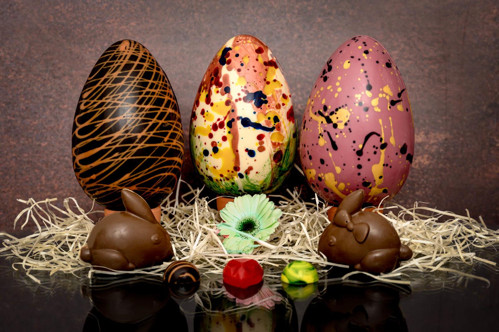 Image shows three chocolate easter eggs side by side on a bed of straw. One is dark chocolate, one white chocolate with splashes of colour and the other is pink with yellow and black marbling. In front of the eggs are two chocolate rabbits with a flower positioned in the middle. In front are three different handmade chocolates, one dark chocolate, one coloured red and the final one is yellow and green.