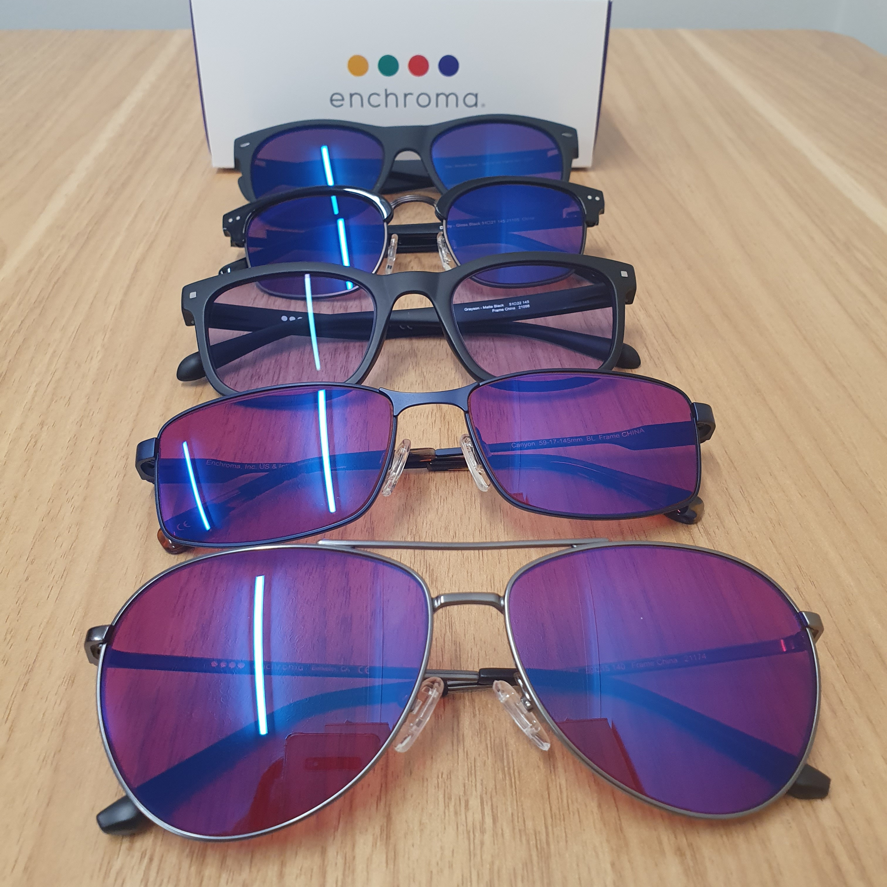 Image of 5 pairs of EnChroma Colour Blindness Glasses in a vertial row with the enchroma presentation box behind them