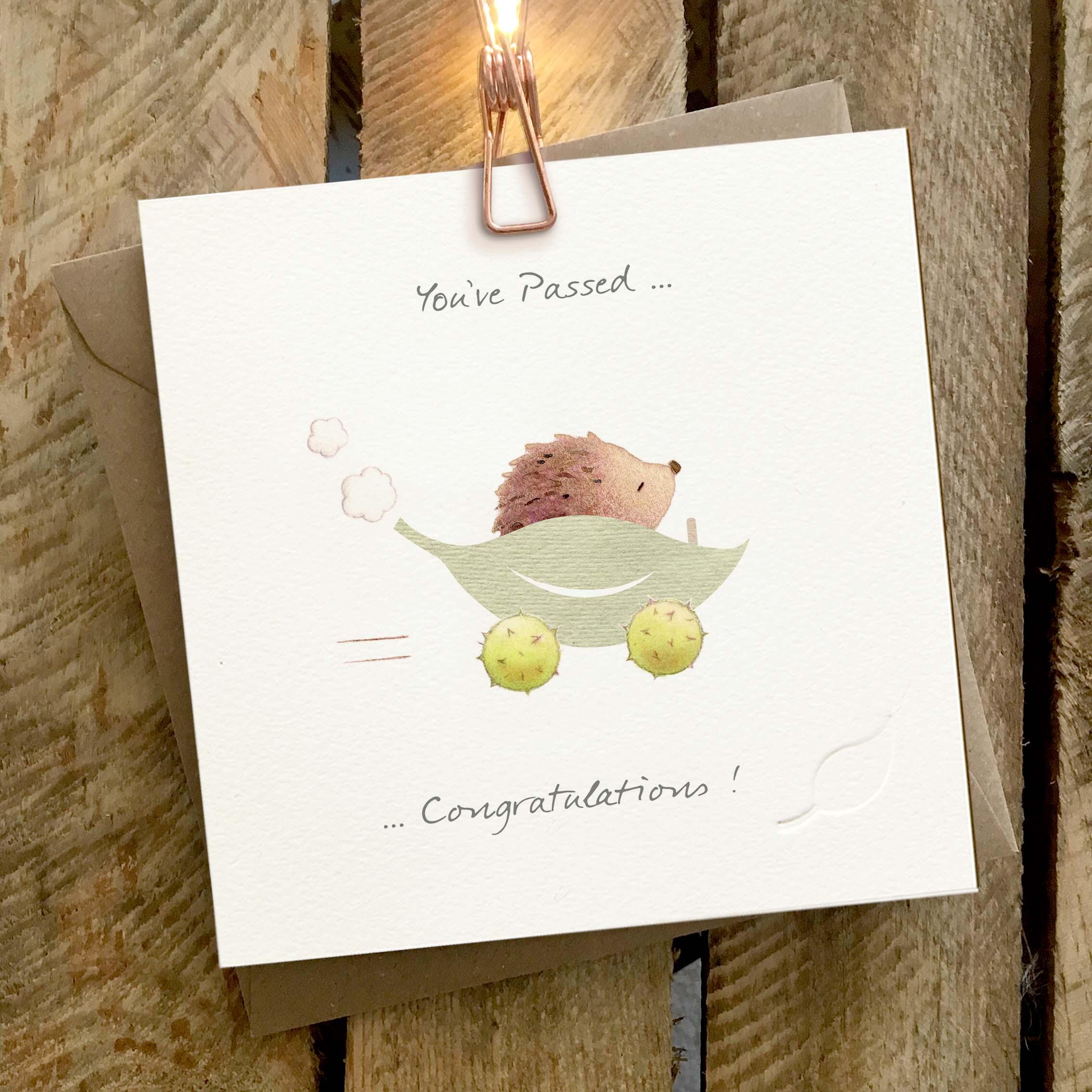 Card featuring a cute hedgehog driving a leaf and conker wheels car. Caption reads ‘You’ve Passed! Congratulations’.