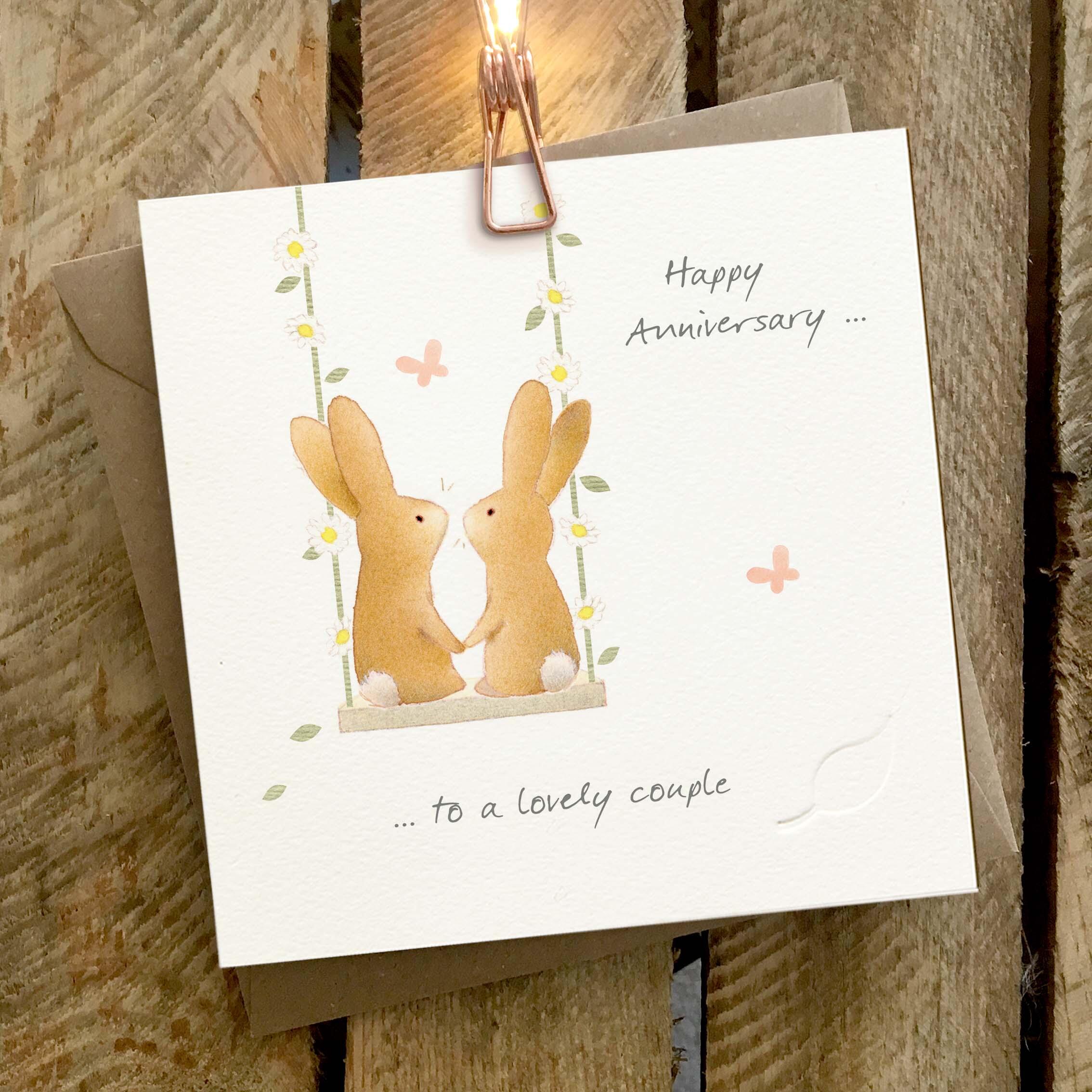 Card featuring a cute rabbit couple sitting on a daisy chain swing. Caption reads ‘Happy Anniversary to a Lovely Couple’.