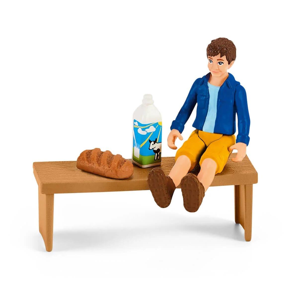 Schleich figure sitting on a bench with bread, milk bottle and food items beside them.