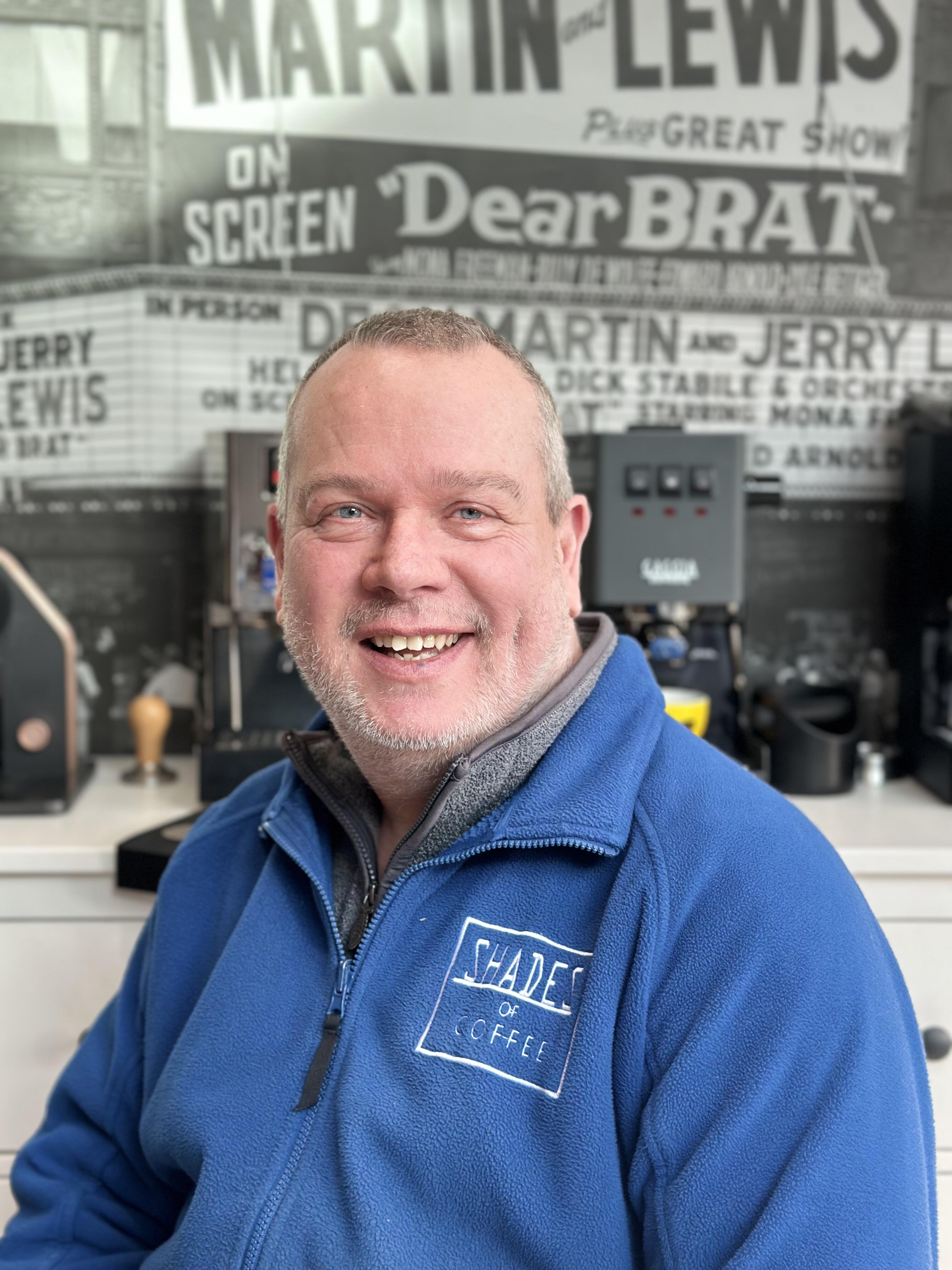 Smiling Shades of Coffee team member wearing a blue branded fleece, sitting in front of a Gaggia coffee machine and vintage poster wall.
