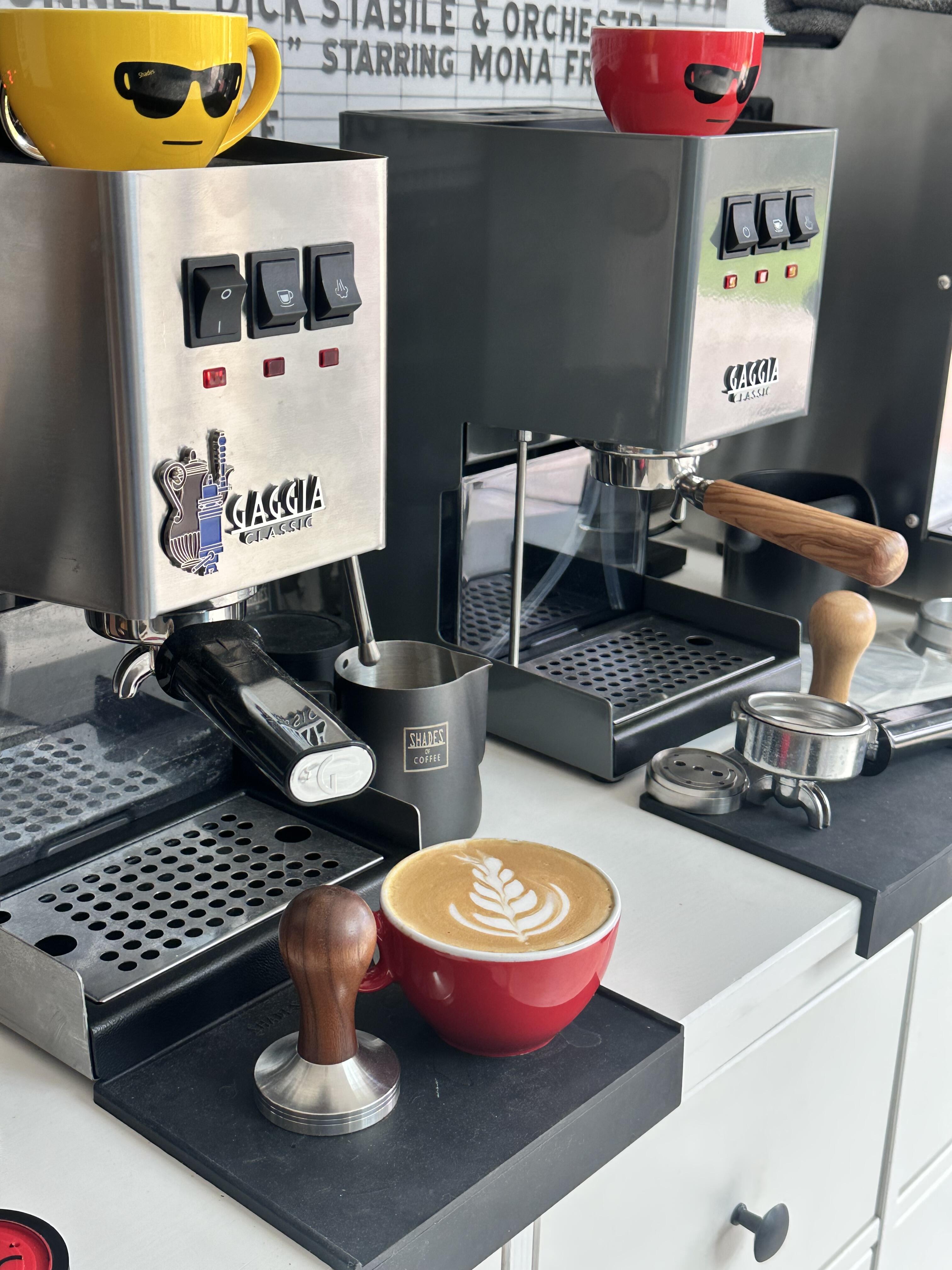 Latte art in a red coffee cup placed beside a tamper, espresso machine, and Shades of Coffee milk frothing jug.