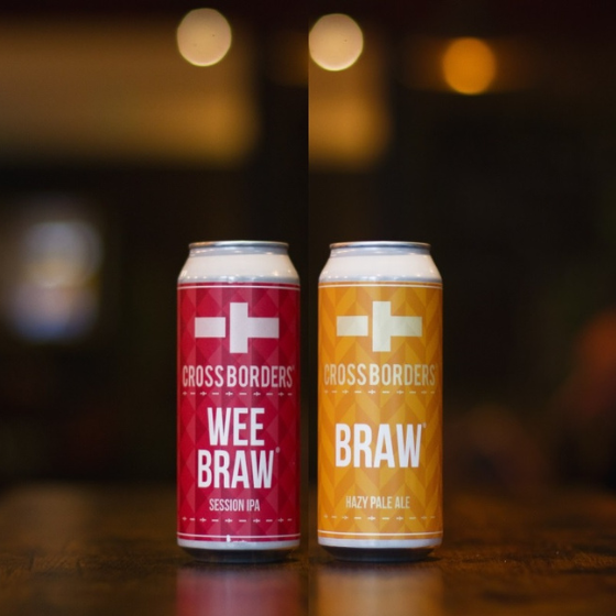 Two cans sit on a dark wooden table. One has a yellow label, Braw, the other has a pink label, Wee Braw.