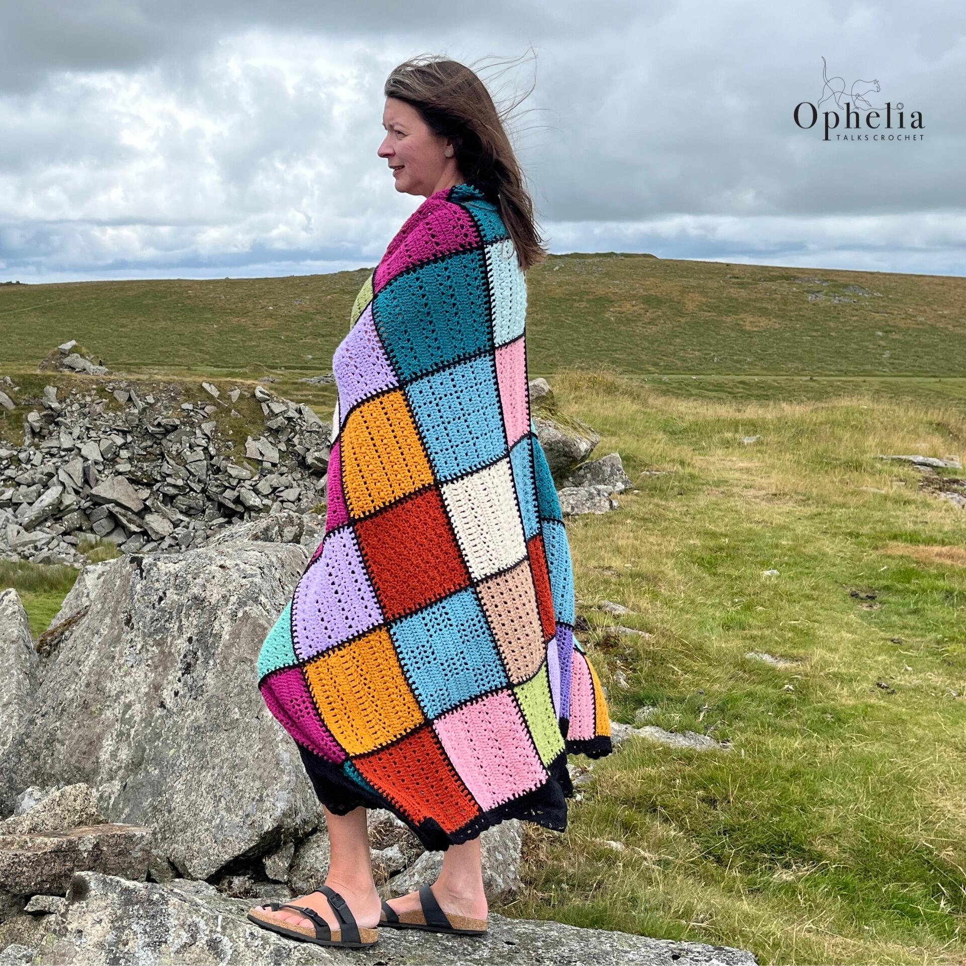 Woman wearing a patchwork crochet blanket standing on rocky terrain near the sea.