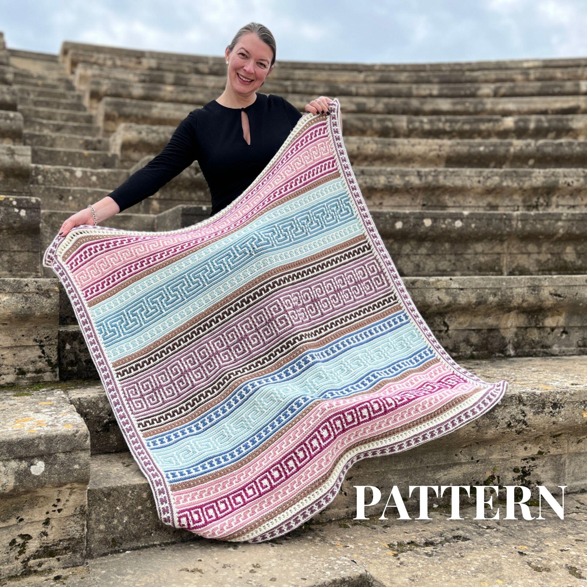 Smiling woman holding a colorful Greek-inspired mosaic crochet blanket with intricate overlay designs in pink, blue, and brown, displayed on ancient stone steps – Odyssey Mosaic Blanket crochet pattern by Ophelia Talks