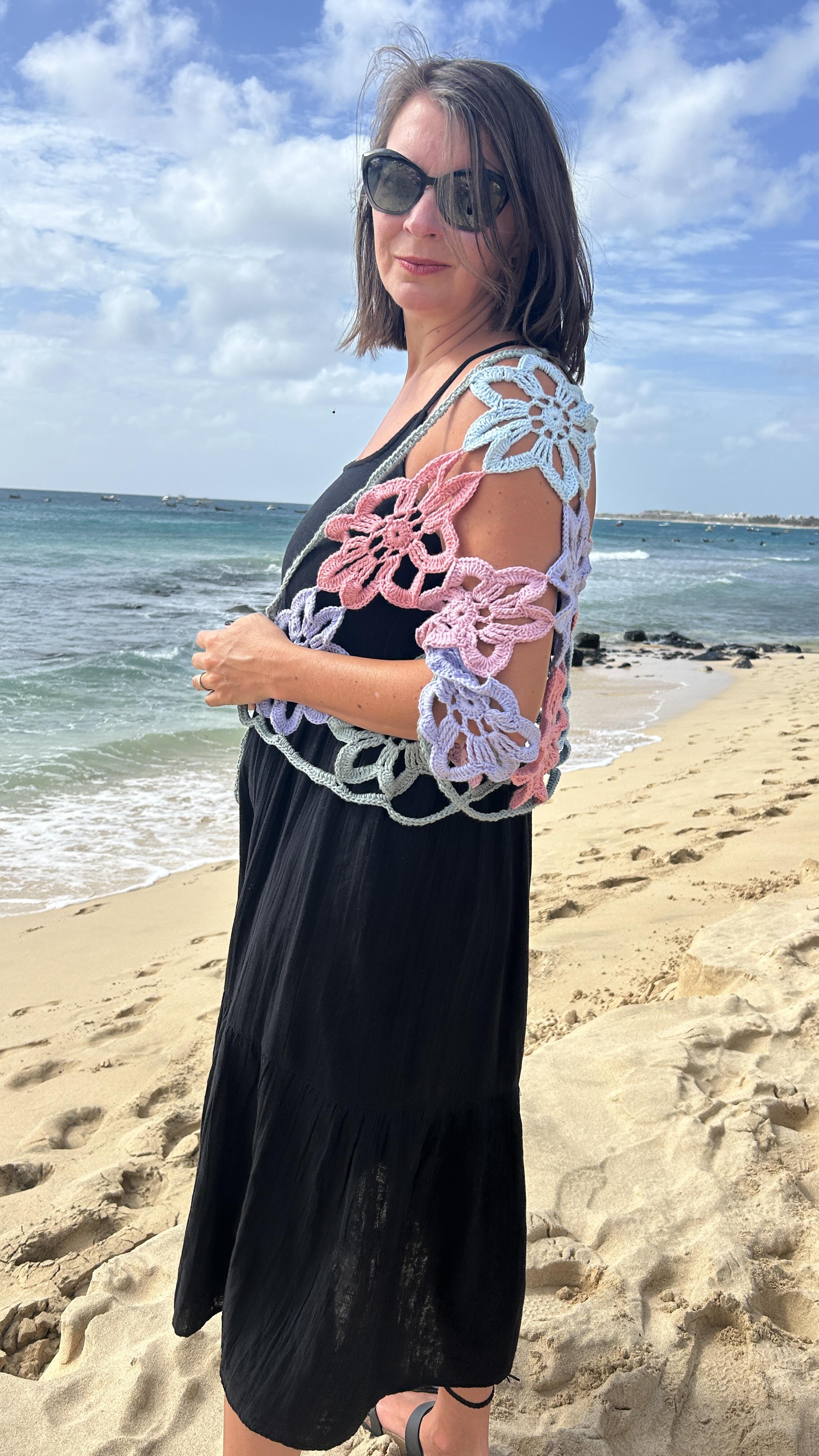 Woman wearing the Flower Power Shawl crochet design on a beach, featuring large pastel crochet flowers in pink, blue, sage and lilac over a black dress.