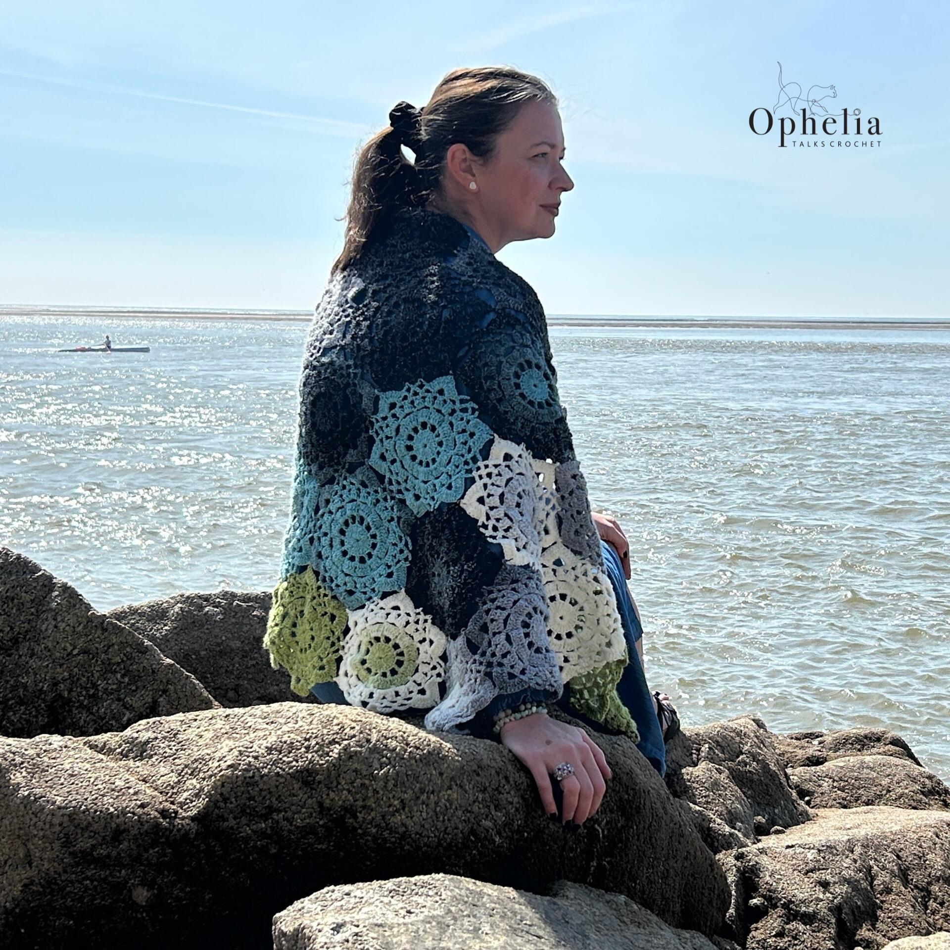 Woman sitting on seaside rocks wearing the Onyx Shawl crochet design made with hexagon motifs in gradient yarn – pattern by Ophelia Talks Crochet