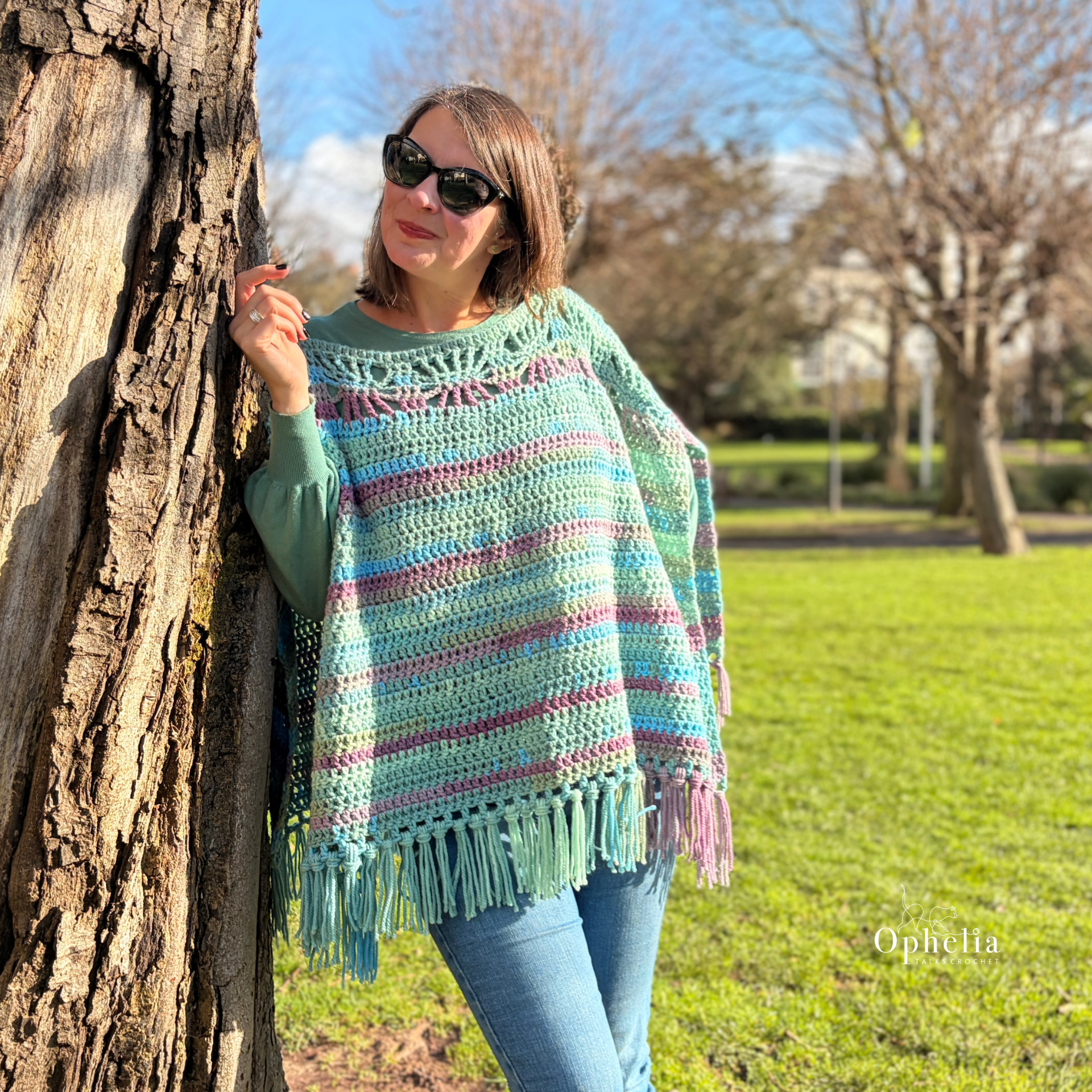 Crochet Garden Slate Poncho worn outdoors in a park, showcasing the ruana style crochet garment with chunky textured stitches, pastel striped yarn and decorative tassel fringe by Ophelia Talks Crochet.
