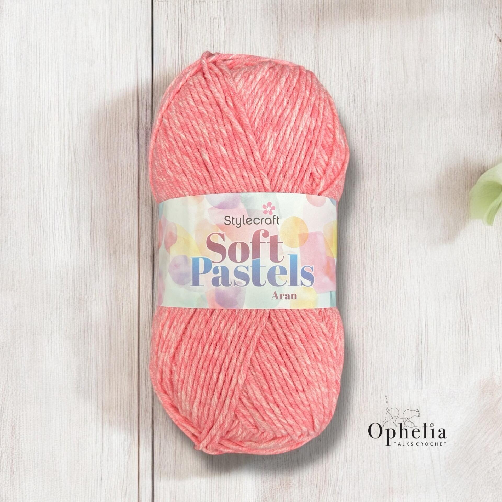 `alt="A skein of Stylecraft Soft Pastels Aran yarn in a soft coral pink shade, resting on a whitewashed wooden background, with the Ophelia Talks Crochet logo in the corner."`