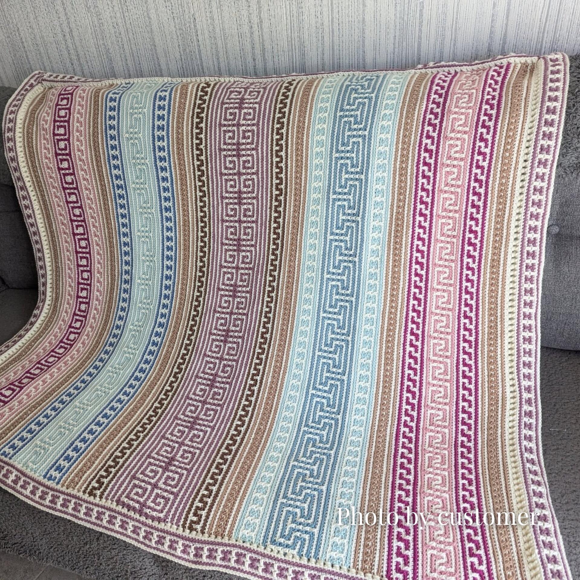 The Odyssey Blanket – a beautifully detailed mosaic crochet design with alternating geometric patterns in soft tones of teal, pink, blue, brown, and cream, draped over a grey sofa. Customer photo.