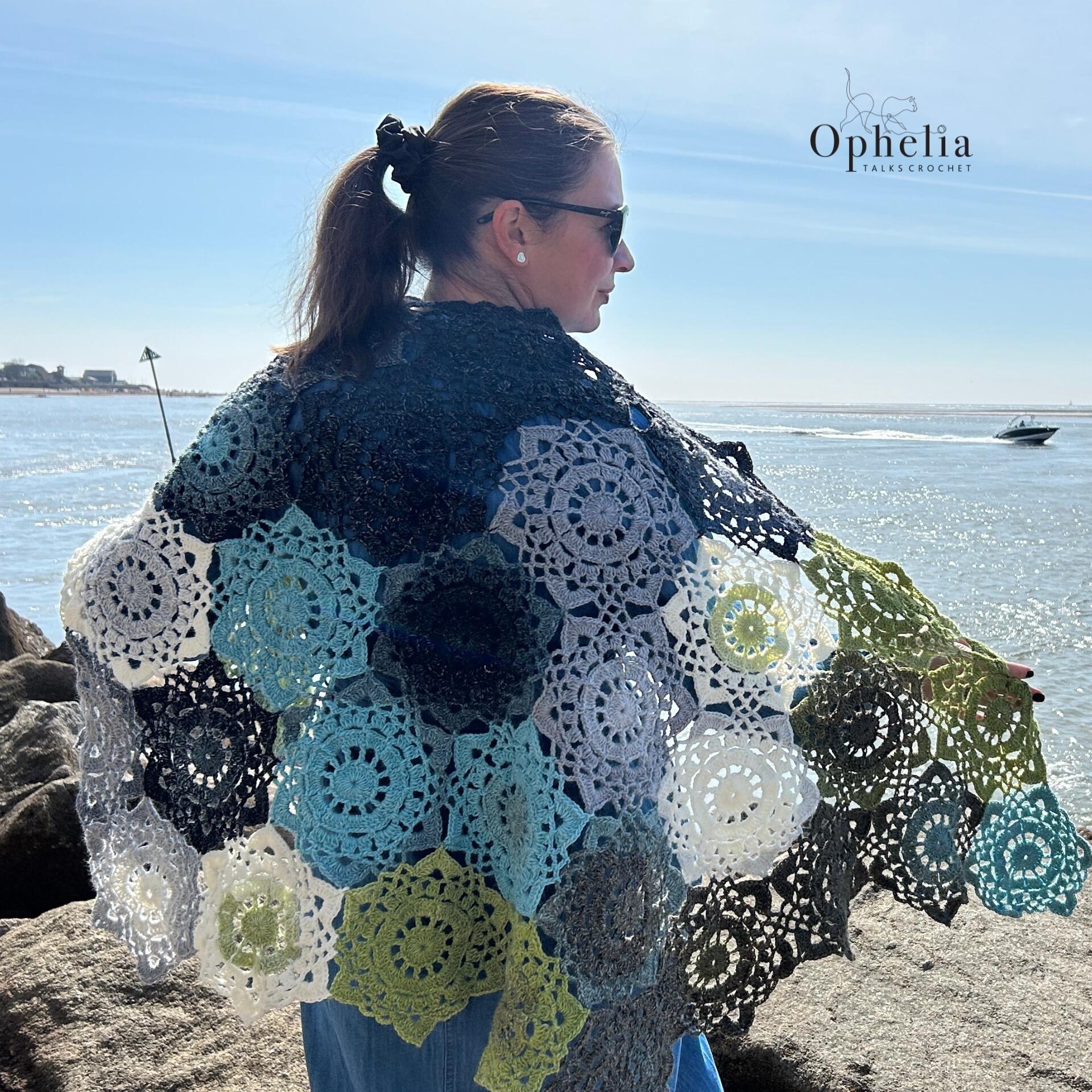 Back view of a woman wearing the Onyx Shawl made from gradient hexagon crochet motifs in blues, greys, and greens, standing on coastal rocks by the sea – Ophelia Talks Crochet.