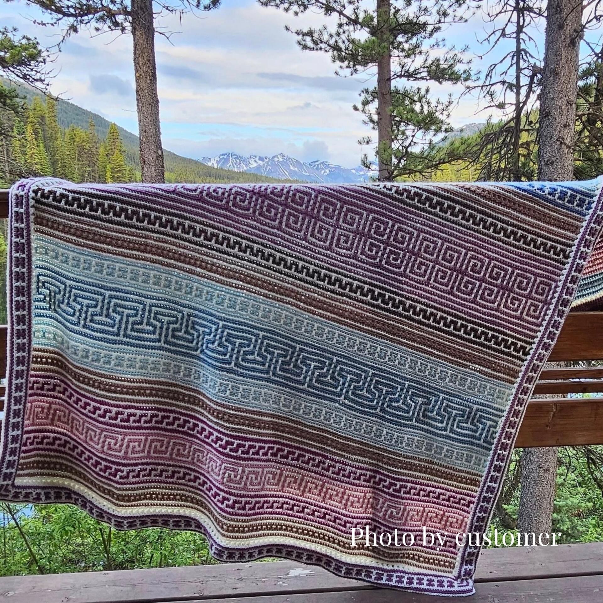 A handmade crochet blanket, the Odyssey Mosaic Blanket, is draped over a wooden railing in an outdoor setting. The blanket features intricate geometric mosaic patterns in rich shades of burgundy, blue, and cream. Behind the blanket is a scenic view of a pine forest, a lake, and distant snow-capped mountains under a partly cloudy sky. Text in the bottom corner reads, “Photo by customer.