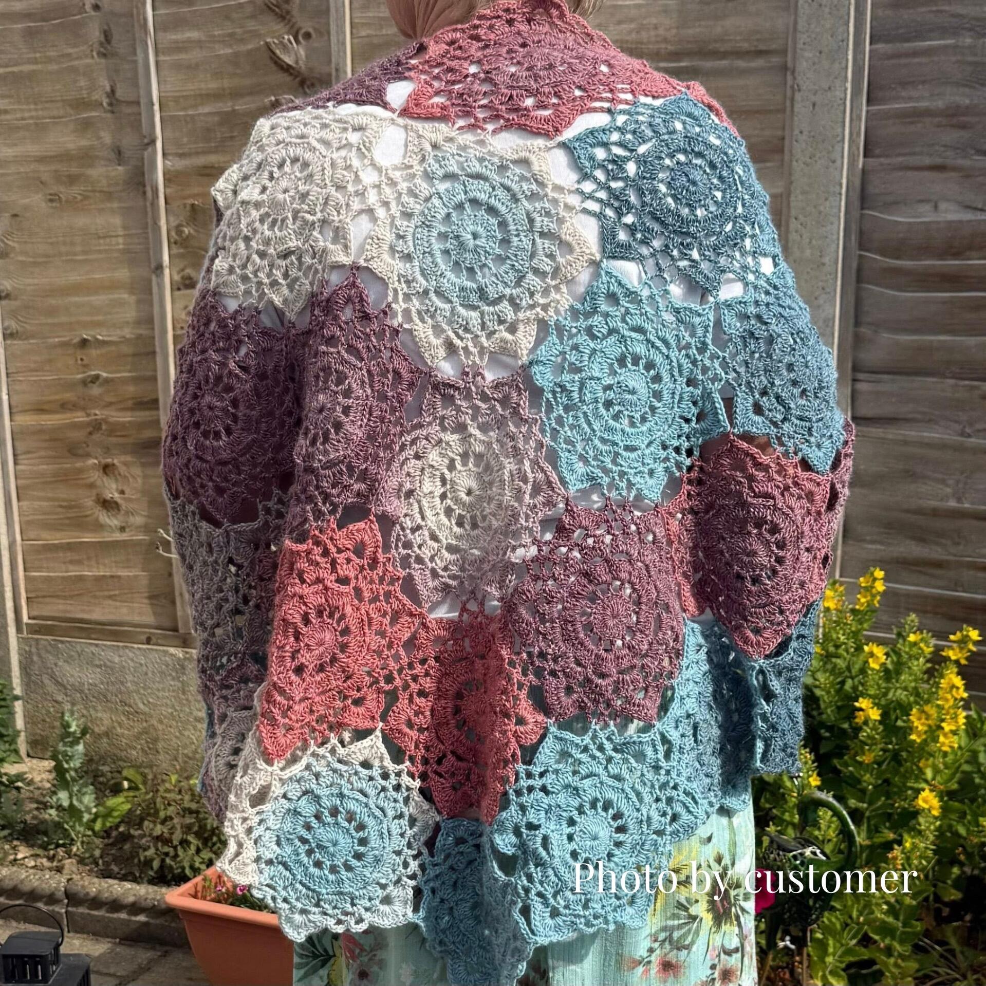 Back view of a person wearing the Onyx crochet shawl made in King Cole Curiosity “Sunset” yarn—a patchwork of lacy circular motifs in muted rose, mauve, teal, and cream—draped over their shoulders against a wooden garden fence.