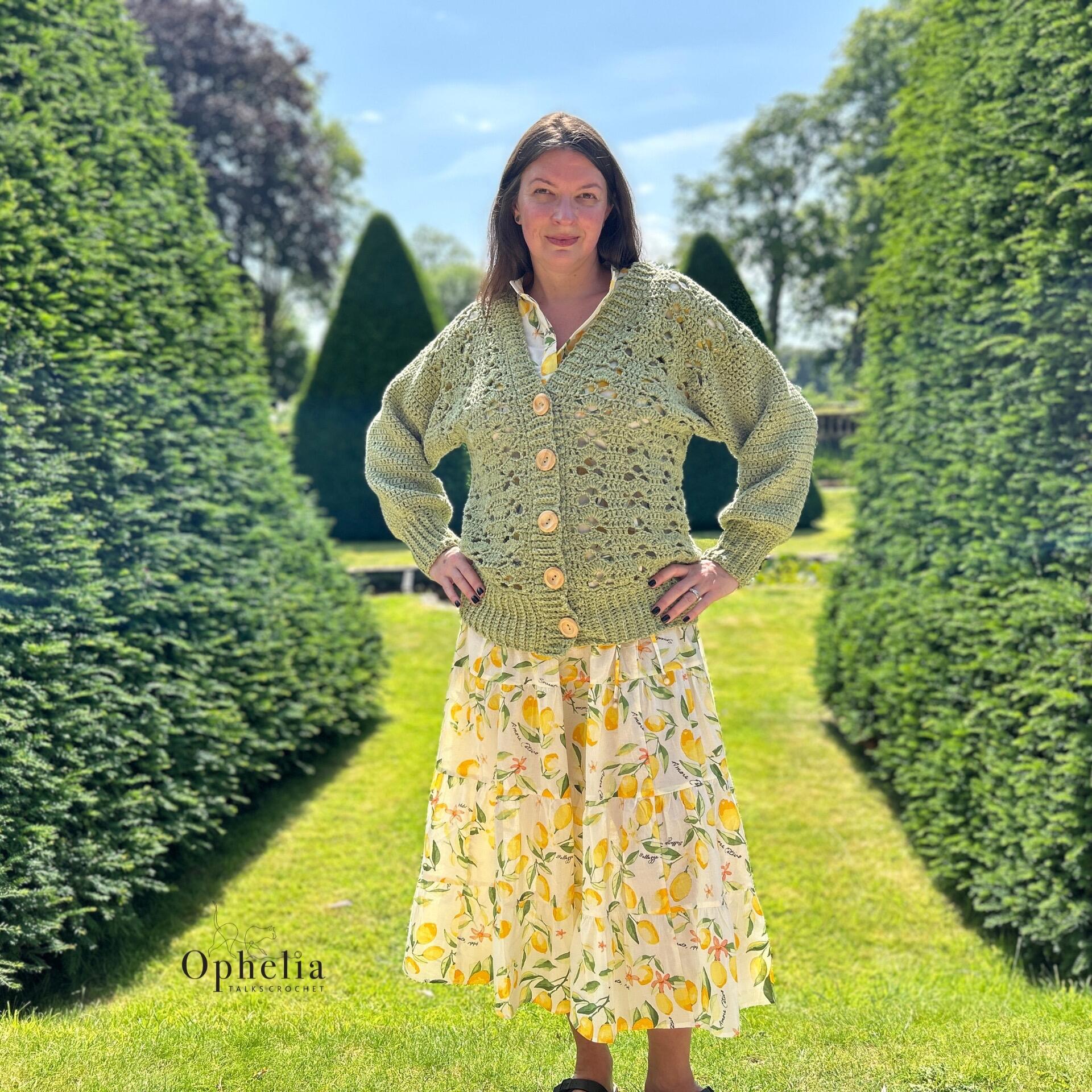 Woman wearing a hand-crocheted Greenwood Cardigan in apple green aran yarn, standing in a sunny garden. The cardigan features balloon sleeves, wooden buttons, and deep ribbing at the cuffs and hem. Designed by Ophelia Talks Crochet.
