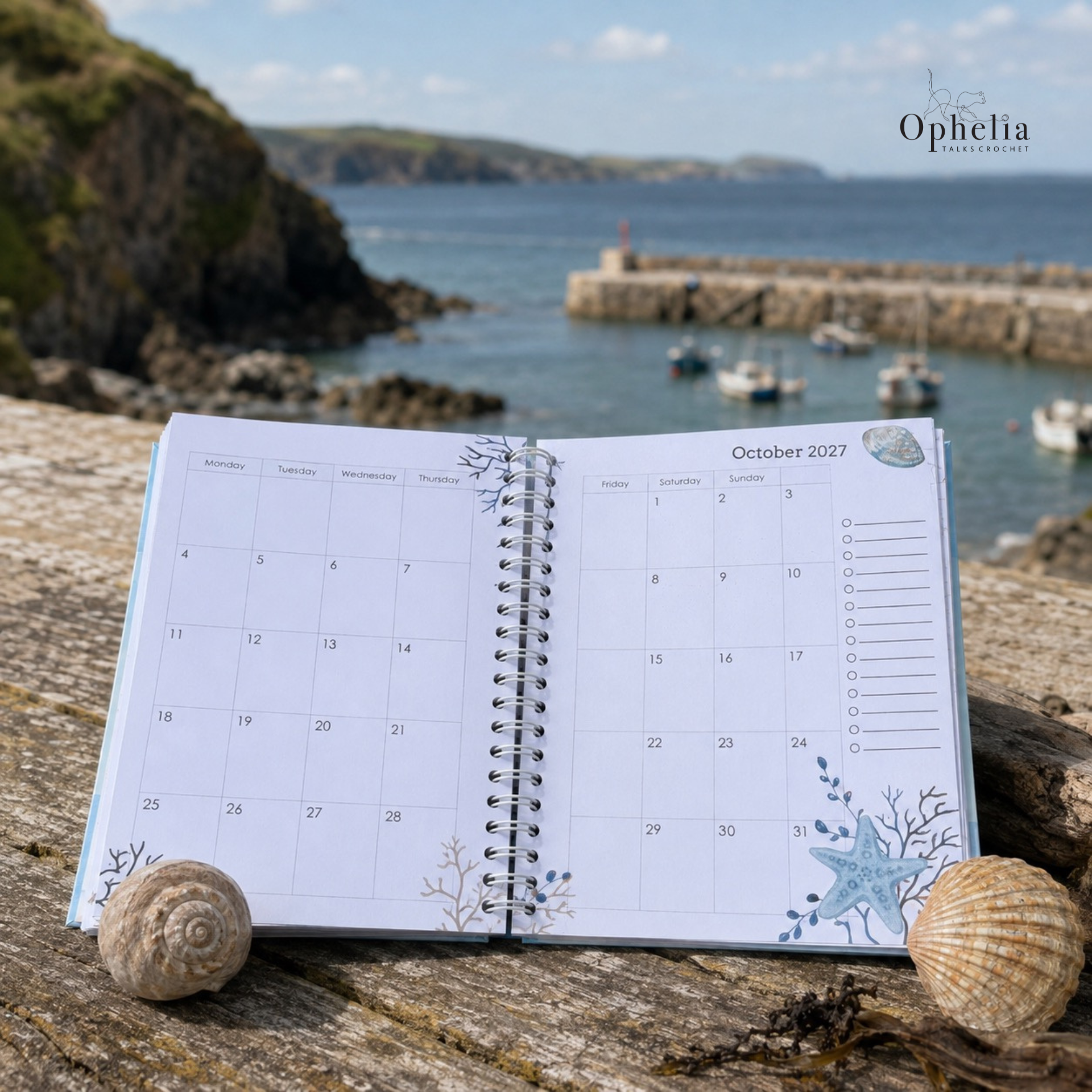 Dream Plan Create Journal open to the October 2027 monthly planner pages, displayed beside seashells with a picturesque Devon harbour and fishing boats in the background.