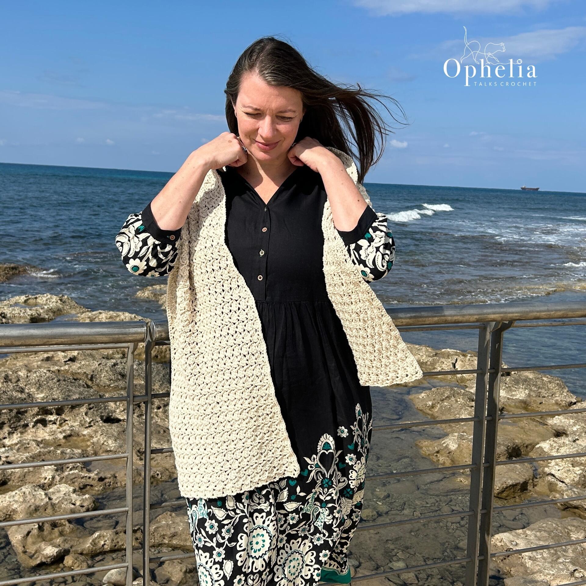 Woman wearing the handmade Persephone Stole crochet wrap in cream cotton yarn, standing by the seaside with waves and rocks in the background. Designed by Ophelia Talks Crochet.