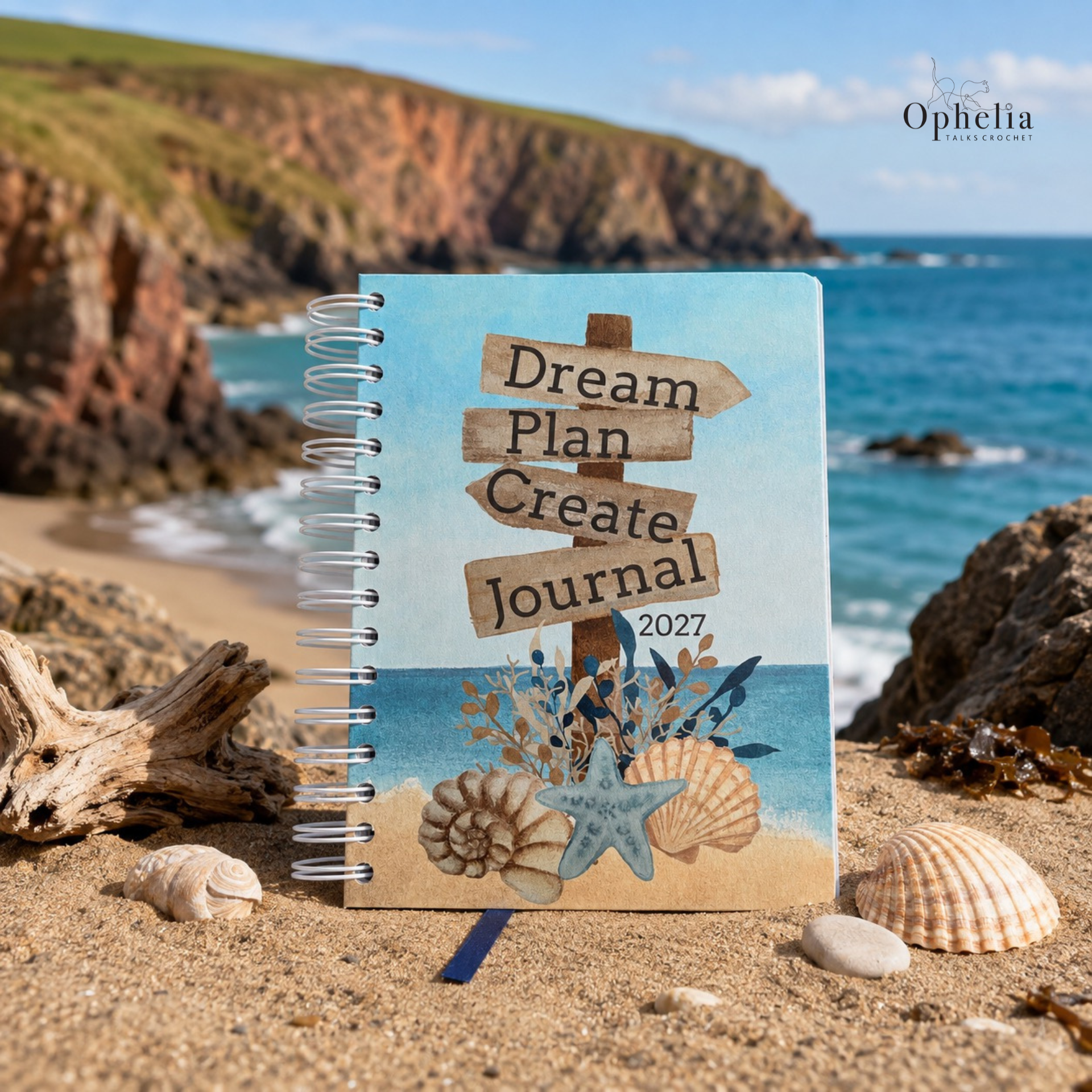 Dream Plan Create Journal 2027 standing on a sandy Devon beach with seashells, driftwood, blue sea and rugged coastal cliffs in the background.