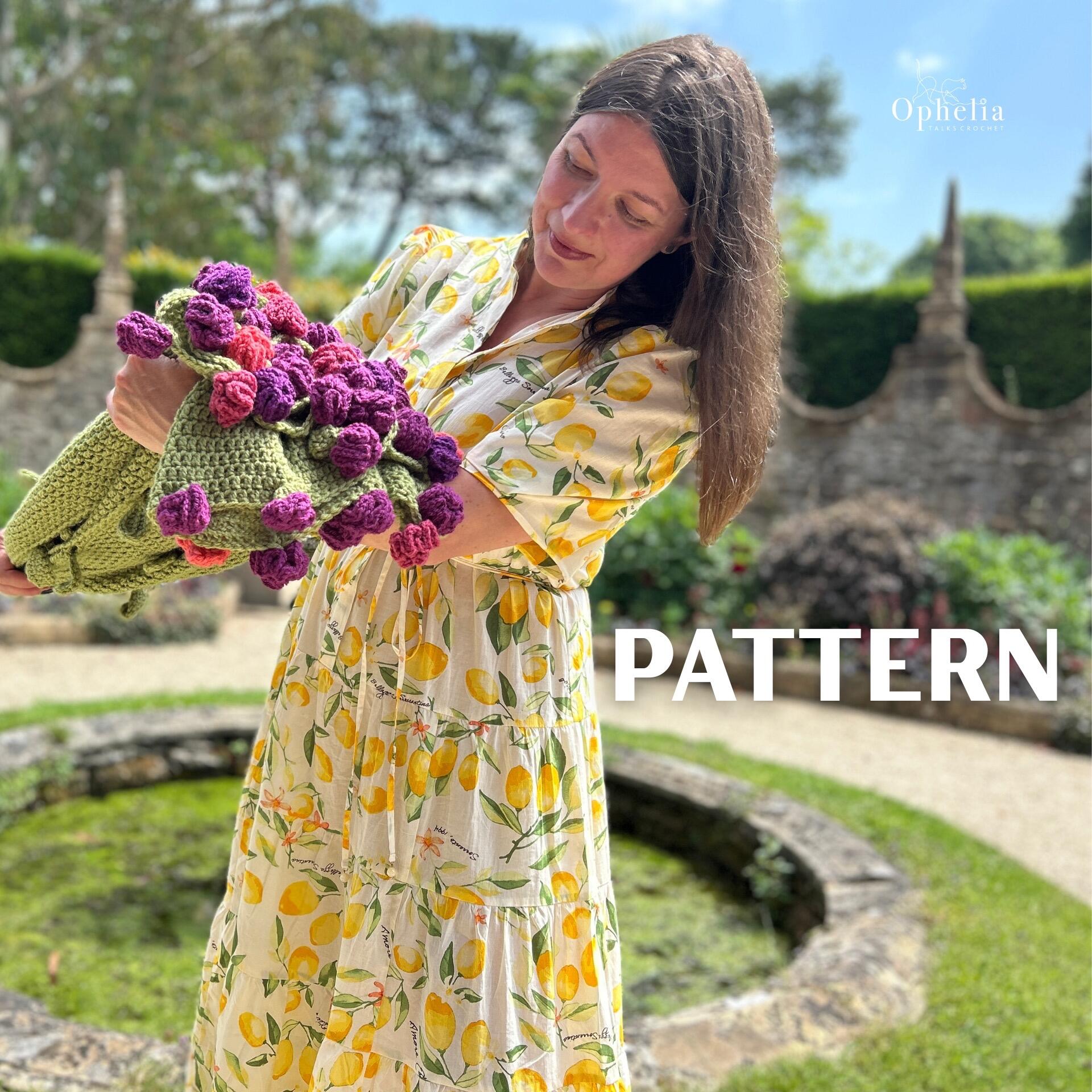 Anja Thys from Ophelia Talks holding the Bouquet Blanket with Carnations, a green round crochet blanket adorned with 3D pink and purple flowers, standing in a sunlit garden wearing a lemon-print dress. The word 'PATTERN' is displayed in bold white text