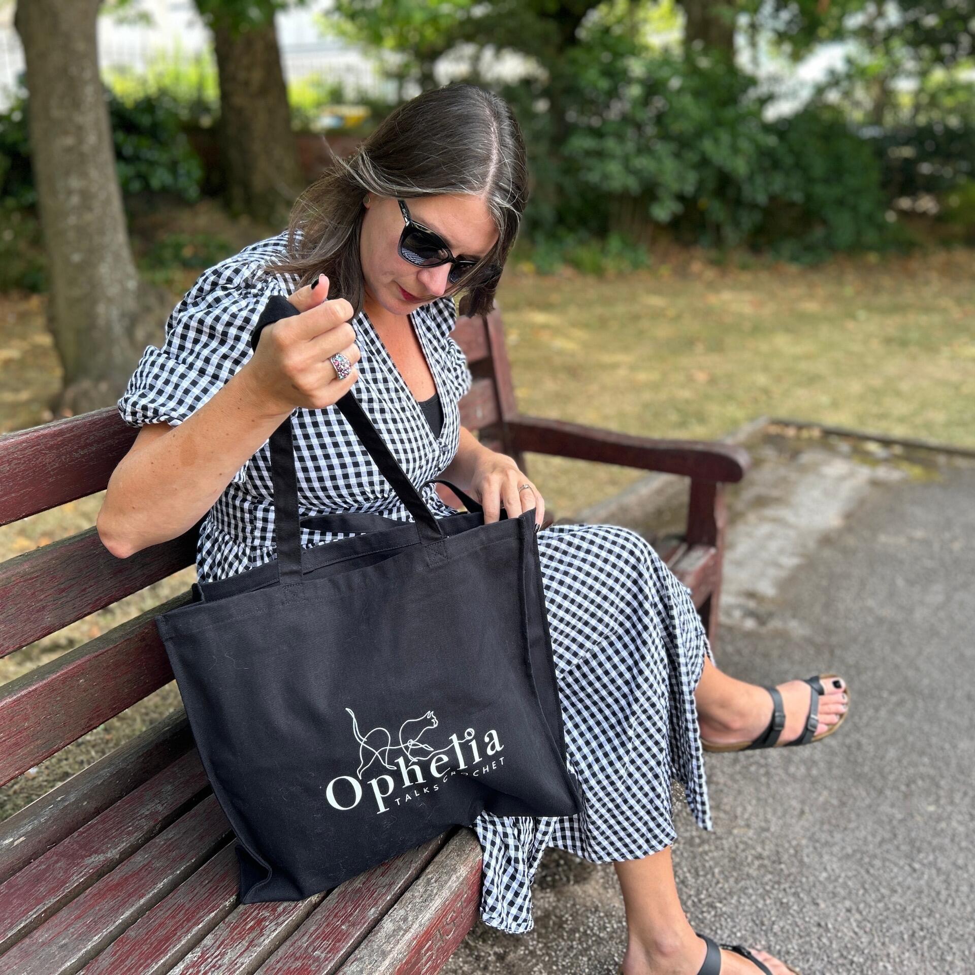 Ophelia Talks Crochet black canvas tote bag with long handles, carried by a woman in a black and white gingham dress while sitting on a park bench, eco-friendly reusable shopper for crochet and everyday use