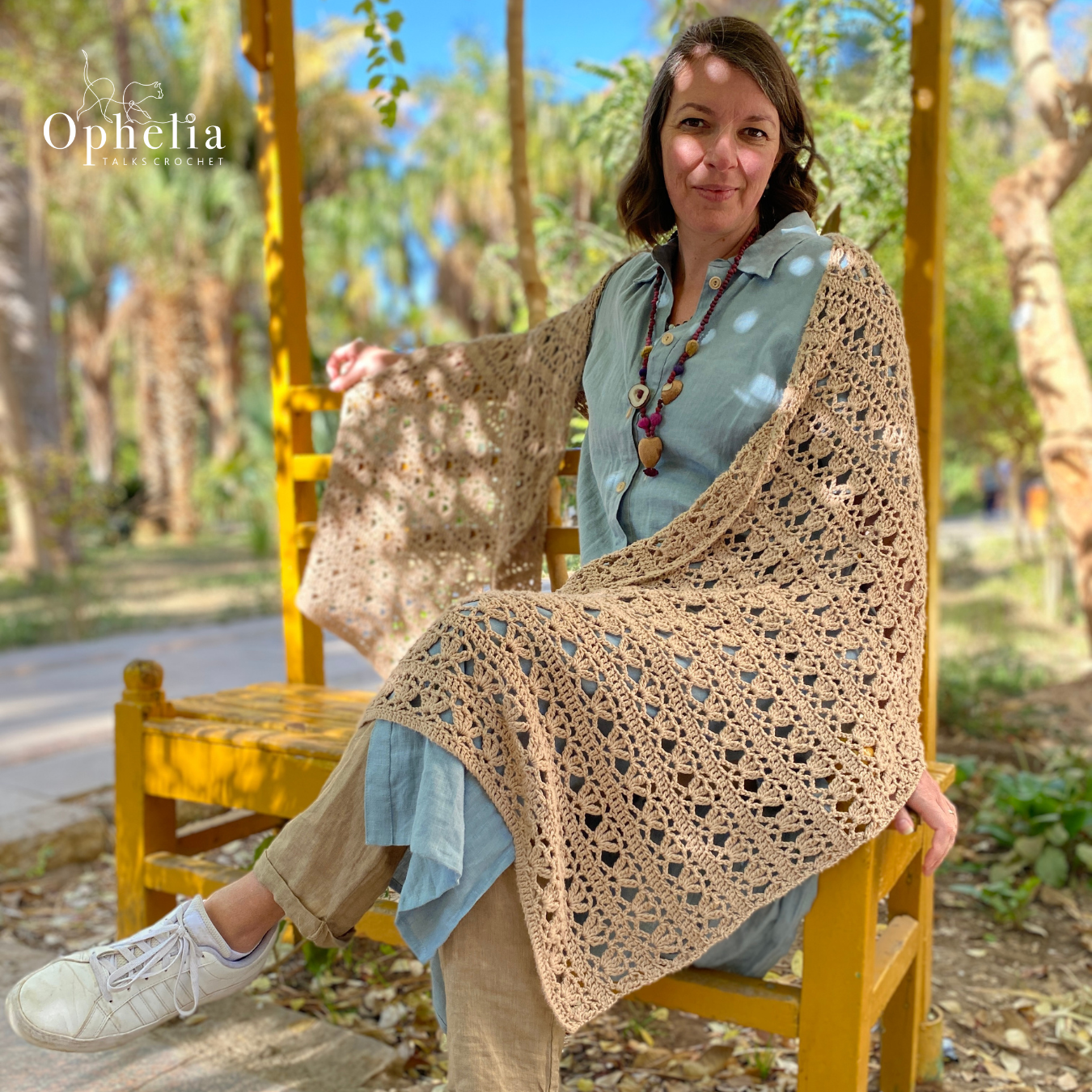 Model wearing a beige, lacy crochet shawl from Ophelia Talks Crochet, seated on a yellow bench in a sunny garden.