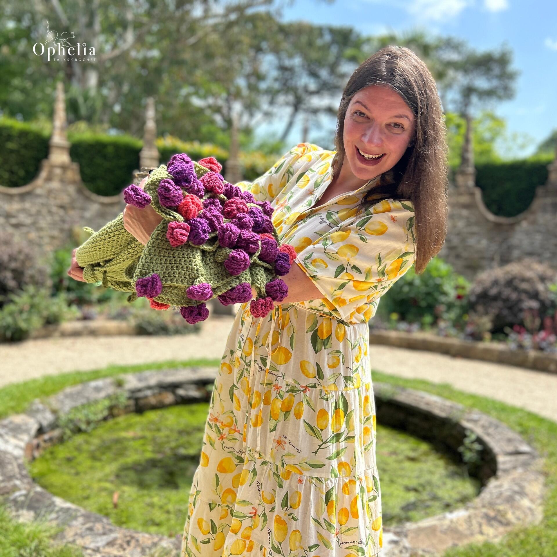 Smiling woman in a lemon-patterned dress holding a green crochet blanket adorned with 3D purple and pink flowers, standing in a sunny garden near a stone pond.