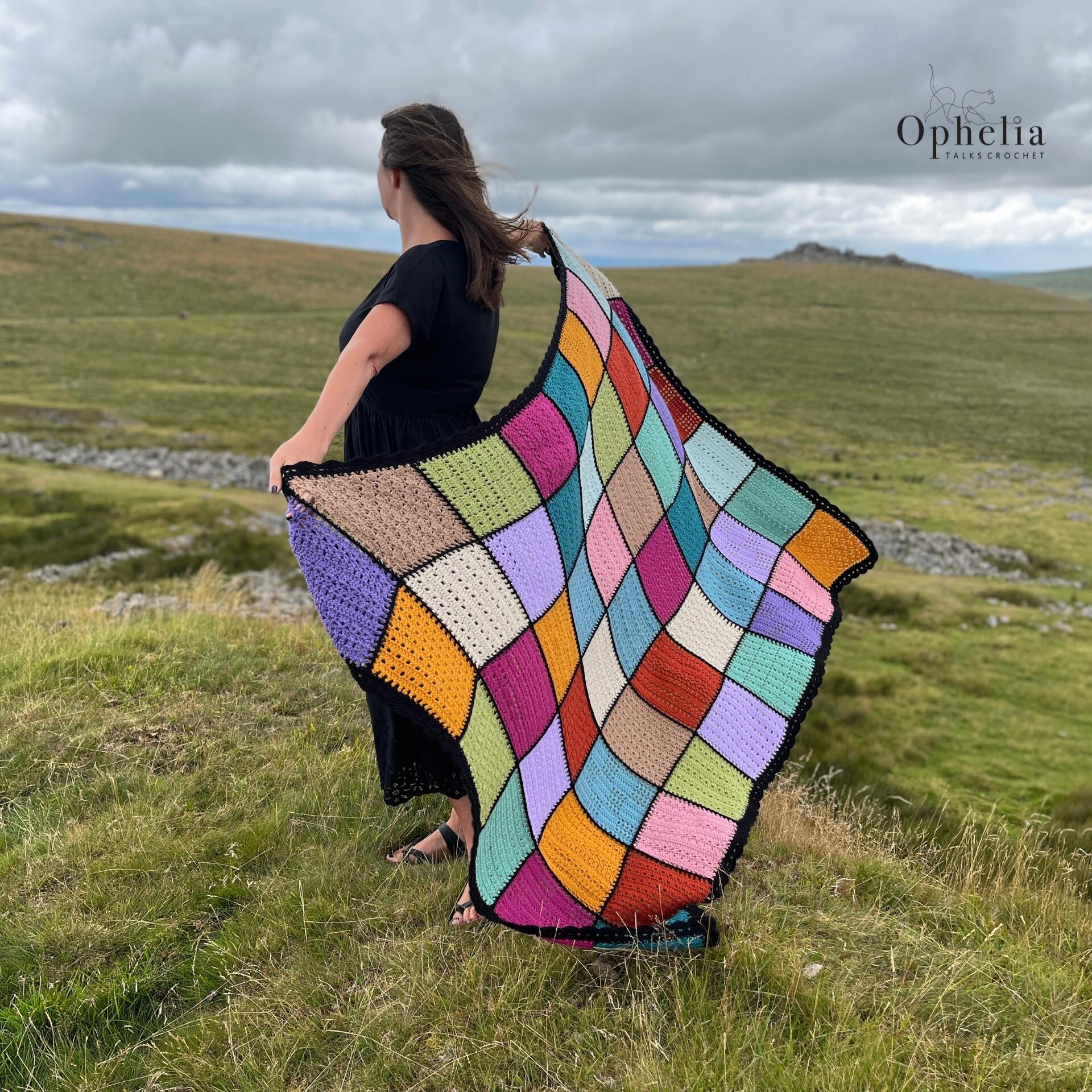 A woman stands on a windswept grassy hill, holding out the Anniversary Blanket by Ophelia Talks Crochet, a colorful, curved patchwork design made to commemorate 10 years of stitches. The blanket ripples in the breeze, showcasing its vibrant colors and black border against a dramatic, cloudy sky and open moorland.