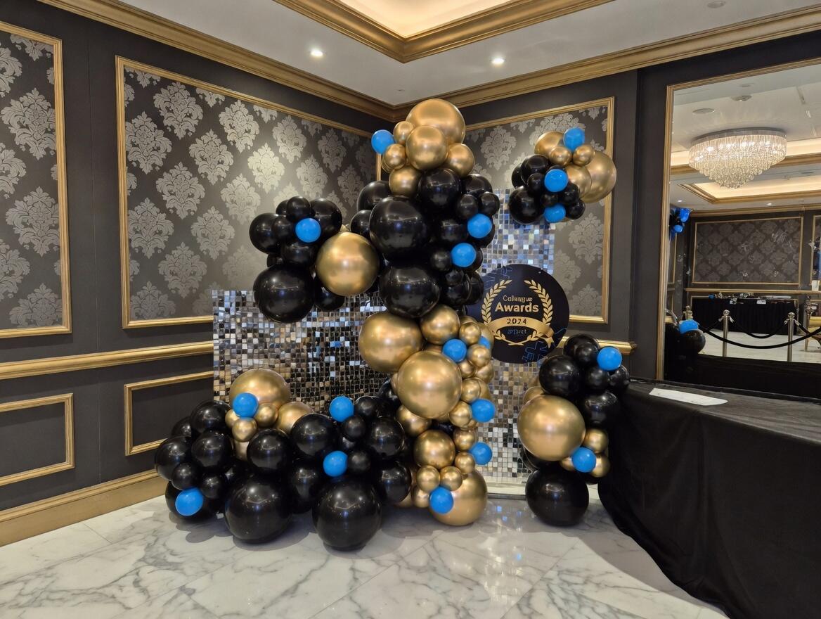 A glamorous balloon decoration for the Colleague Awards 2024, featuring a large organic balloon arrangement in black, gold, and blue. The display stands in an elegant event hall with dark walls, gold trim, marble flooring, and a sparkling mosaic backdrop with the official awards sign.
