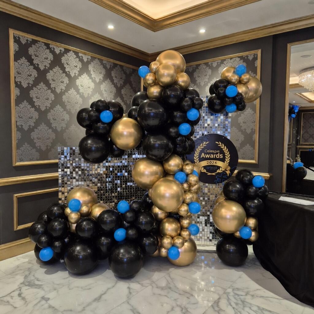 A glamorous balloon decoration for the Colleague Awards 2024, featuring a large organic balloon arrangement in black, gold, and blue. The display stands in an elegant event hall with dark walls, gold trim, marble flooring, and a sparkling mosaic backdrop with the official awards sign.