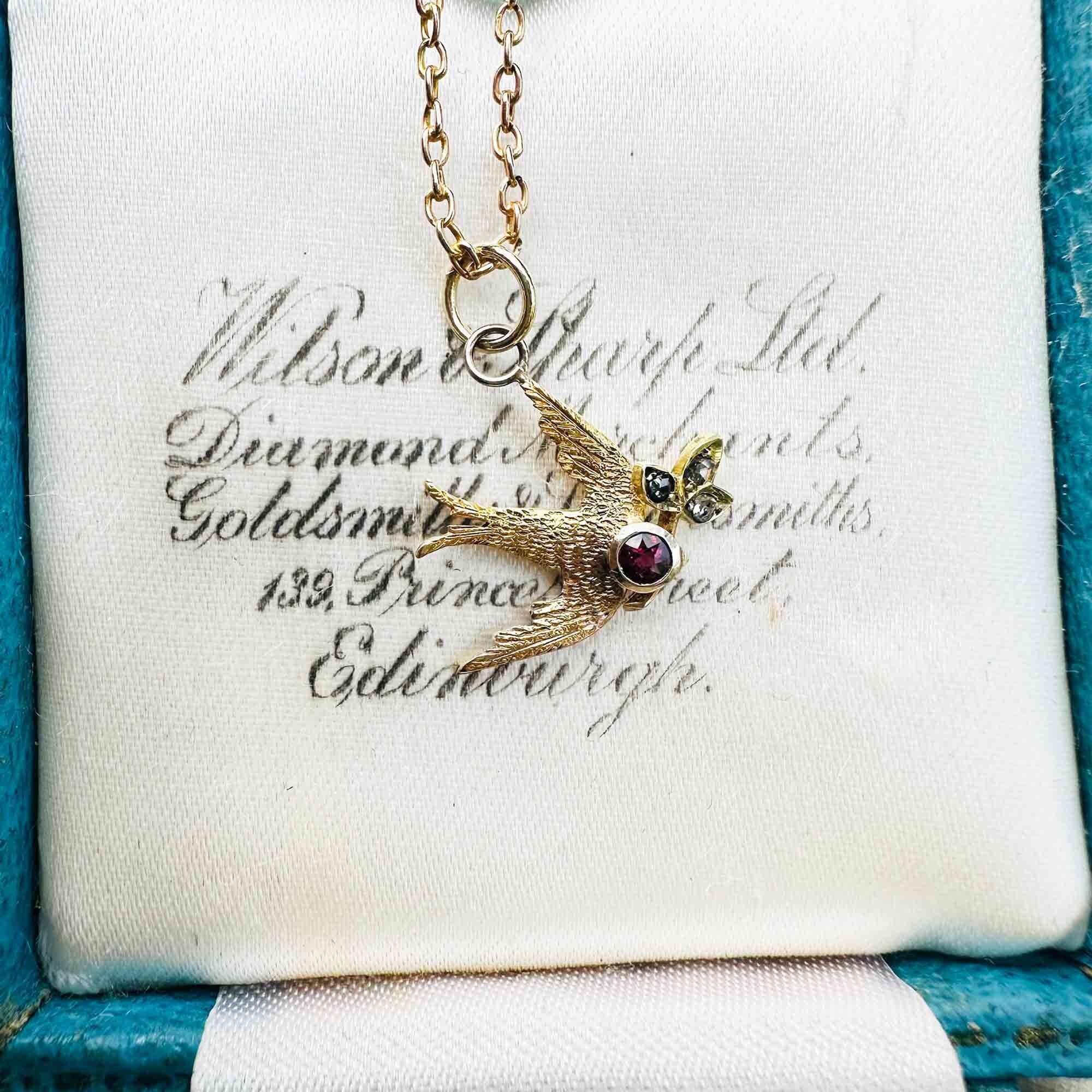 Art Nouveau 15k gold swallow pendant with outstretched wings, holding a diamond trefoil twig in its beak and set with a tiny ruby at the head.