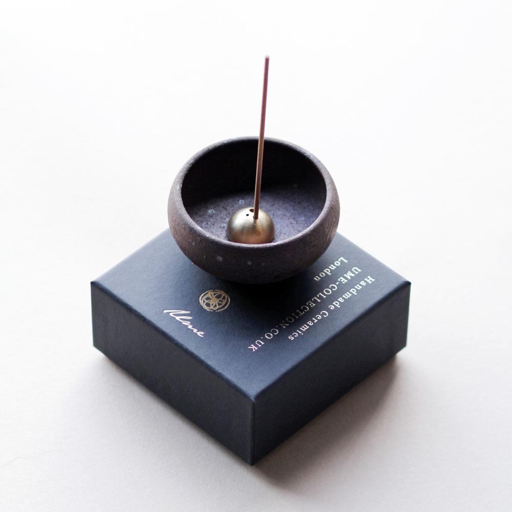 A small black clay bowl shown with brass dome incense holder inside with incense stick inserted on top of a black square gift box.