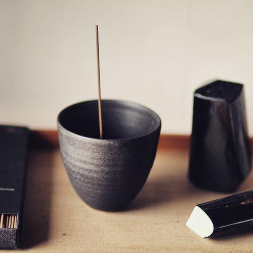 Black porcelain curved pot shown with on a wooden table top with black boxes of incense