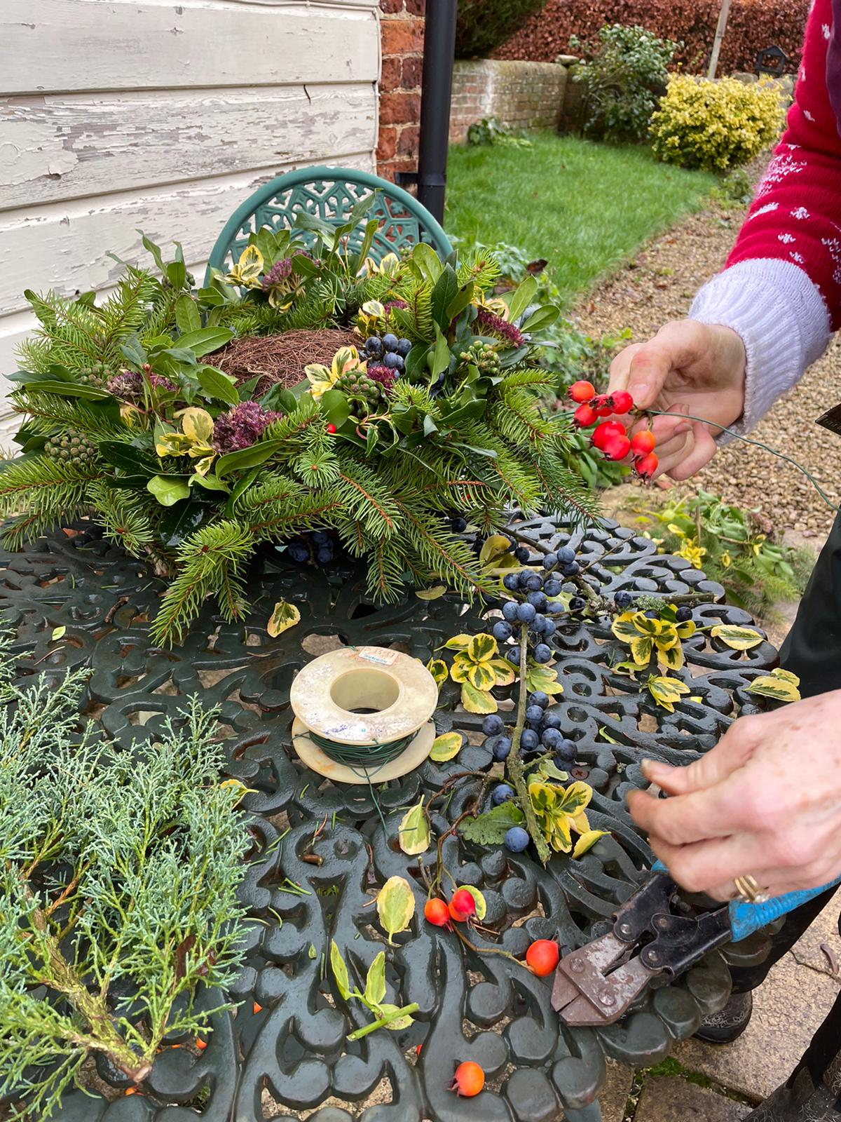 A womans hand is shown decorating a wreath with green foliage on a green wrought iron round garden table