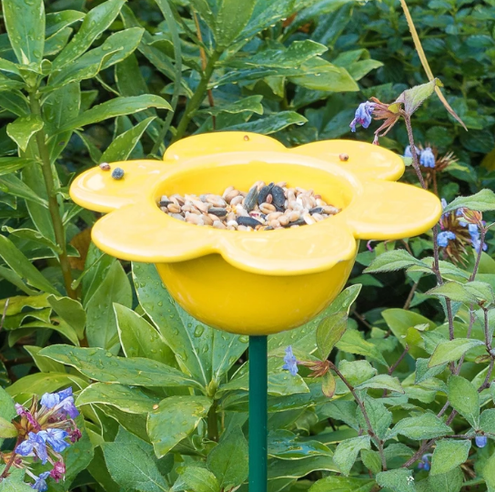 yellow ceramic flower bird feeder on a green metal pole. Shown in a garden background.