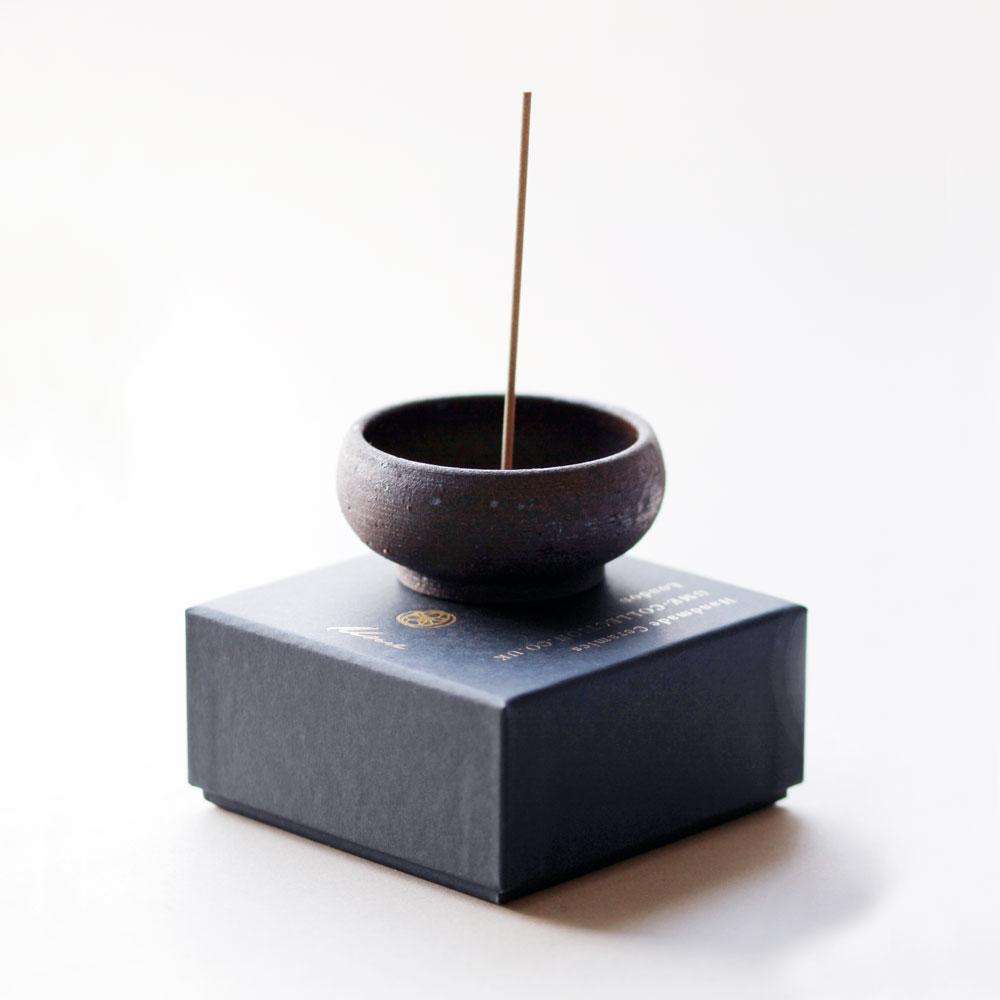 Side view of a small black clay bowl on top of a black gift box with incense stick inserted.